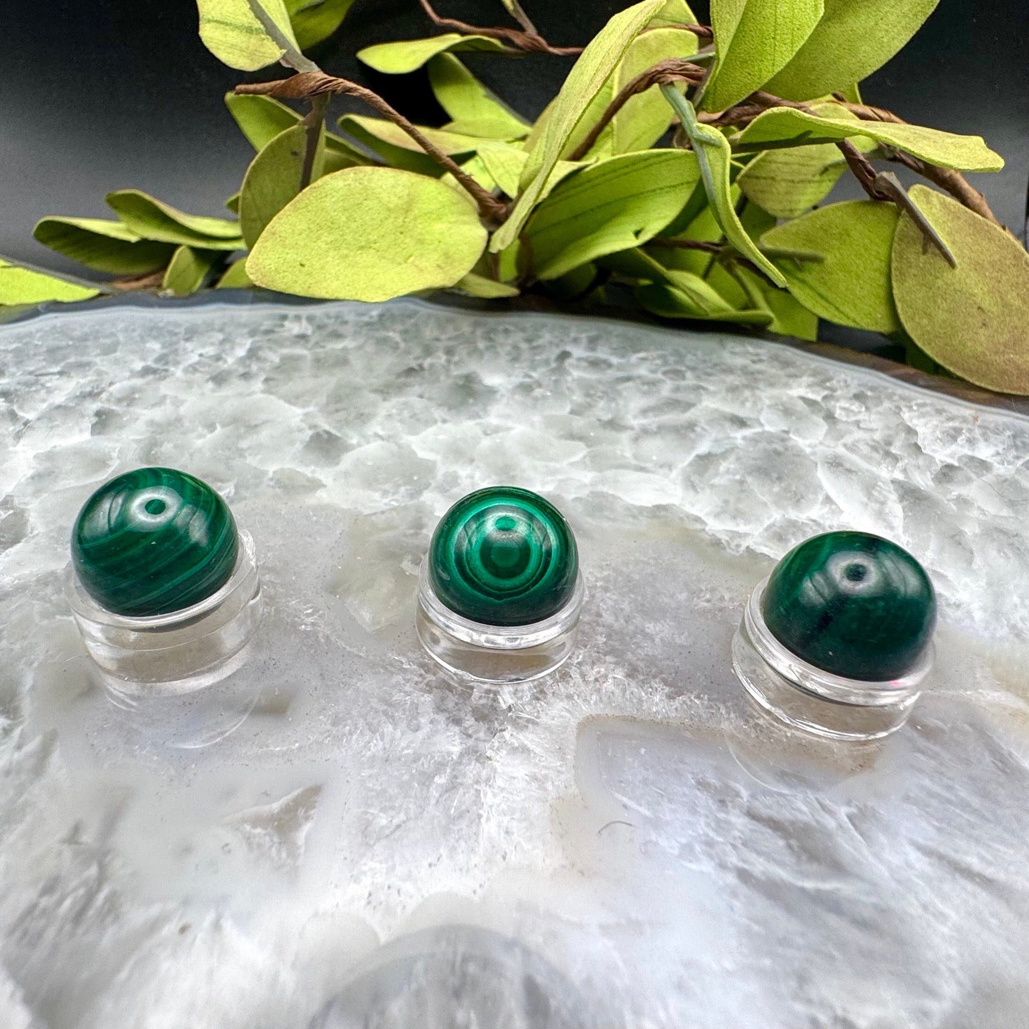 Three green gemstones in clear holders on a textured white surface with green leaves in the background.