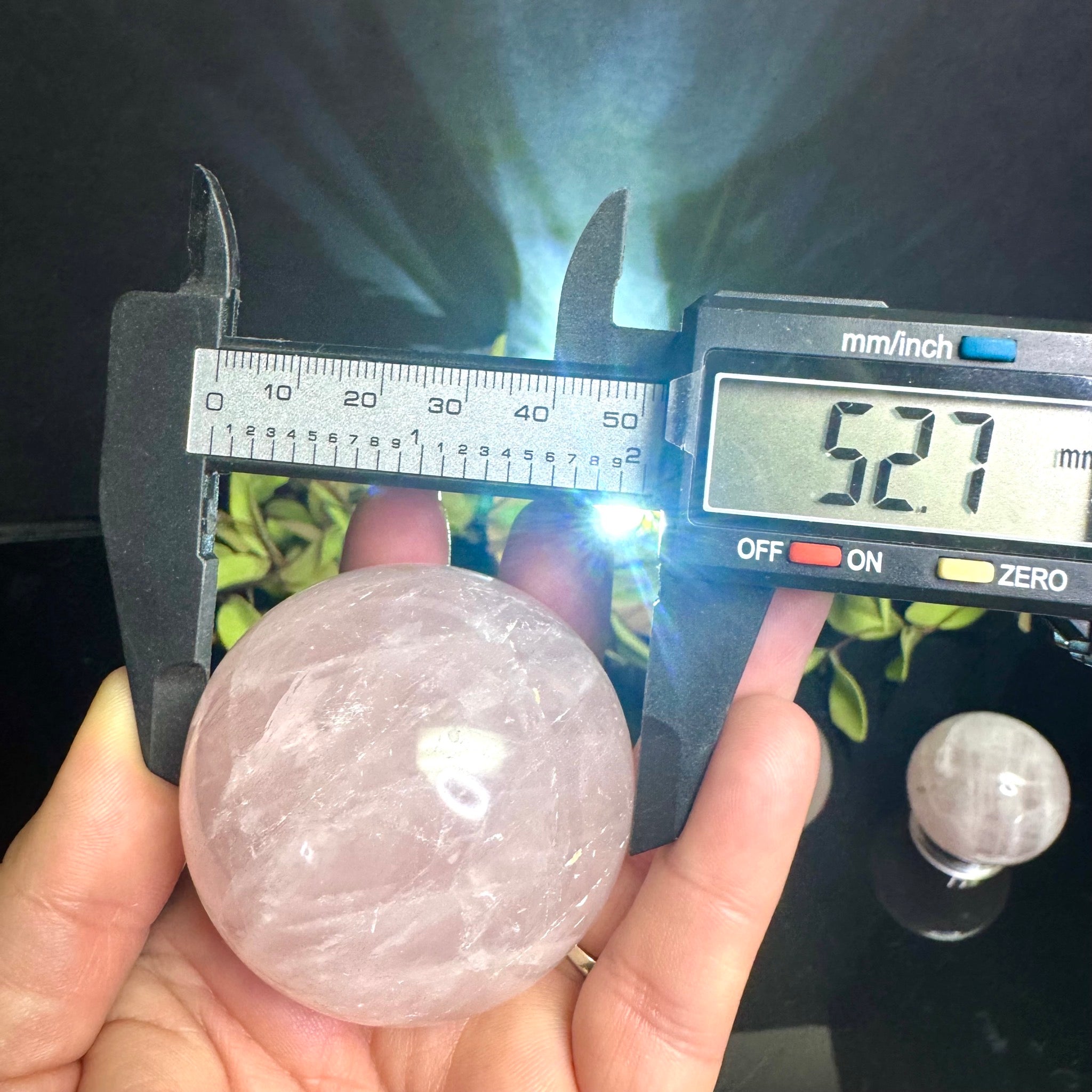Digital caliper measuring a Rose Quartz crystal sphere held by a hand.